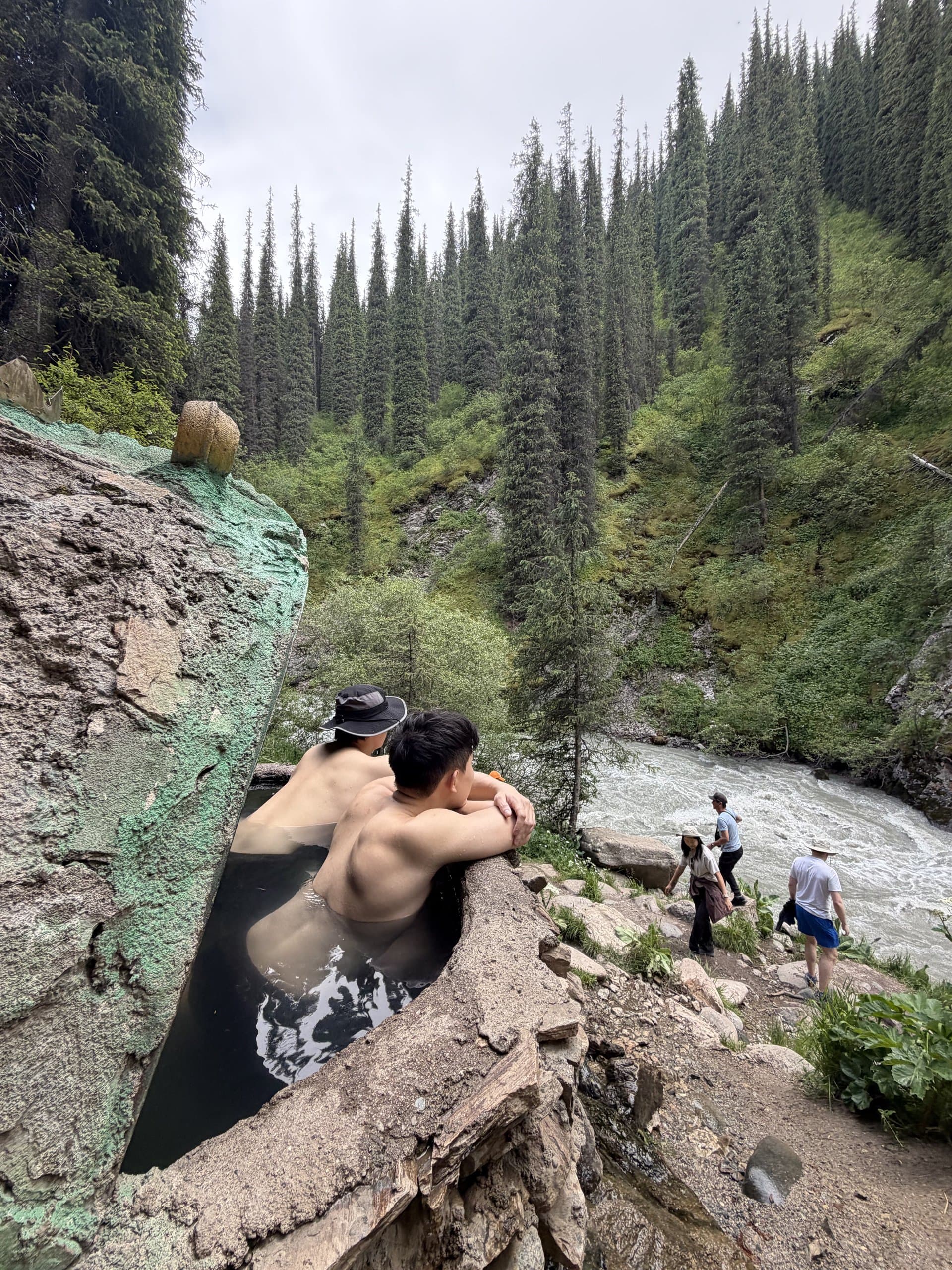 A traveller soaking in a natural hot spring pool beside a mountain river surrounded by spruce forest
